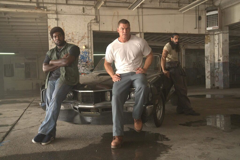 Alan Ritchson leans against a muscle car alongside two crew members in a stripped-down garage in "Motor City." - The Latino Slant